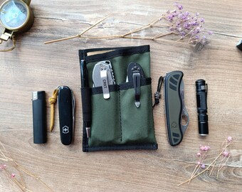 May include: An olive green nylon pocket organizer with two vertical slots containing two folding knives with metal clips. A black pen is attached to the side. Surrounding items include a compass, a black lighter, a black multi-tool, a green folding knife, and a small black flashlight.