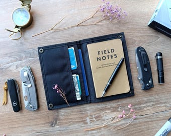May include: A black leather wallet with multiple pockets and slots for cards and cash. The wallet is open to reveal a notebook with the words "Field Notes" on the cover. A pen is also visible in the wallet.