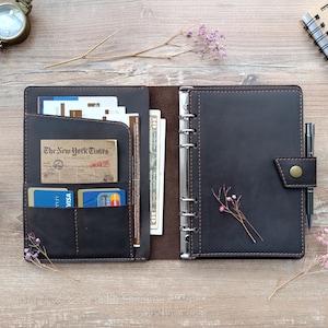 May include: A dark brown leather planner with a six-ring metal binder mechanism, a snap button closure, and a pen loop. The interior features multiple pockets containing credit cards, paper documents, and a ten-dollar bill.