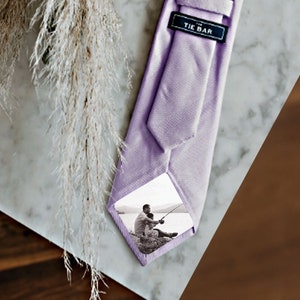 May include: A lavender tie with a white photograph of a person fishing on the front. The tie has a black label that says "The Tie Bar" on it.