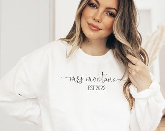May include: White sweatshirt with the text "mrs montana" and "EST 2022" in a black script font. The sweatshirt is a crew neck style and is made of a soft material. The model is wearing blue jeans.