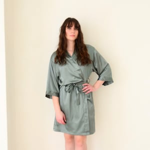 May include: A sage green satin robe with a self-tie sash. The robe has long sleeves and a relaxed fit.