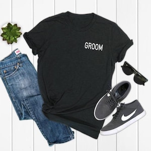 May include: A black t-shirt with the word "Groom" printed on the front pocket. The shirt is laid out on a white wooden surface with a pair of jeans, sunglasses, and sneakers.