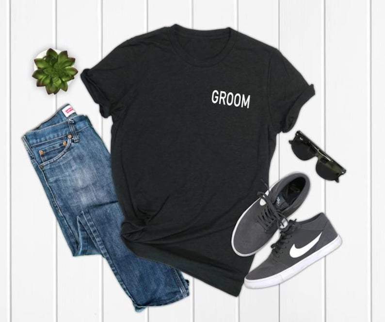 May include: A black t-shirt with the word "Groom" printed on the front pocket. The shirt is laid out on a white wooden surface with a pair of jeans, sunglasses, and sneakers.