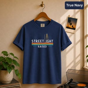May include: A true navy blue short-sleeve t-shirt featuring a graphic print with the text STREET LIGHT RAISED. The design includes a stylized street lamp icon above the text and a series of horizontal stripes in orange, yellow, and teal colors.
