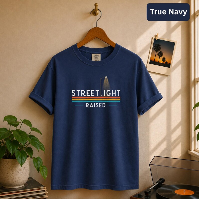 May include: A true navy blue short-sleeve t-shirt featuring a graphic print with the text STREET LIGHT RAISED. The design includes a stylized street lamp icon above the text and a series of horizontal stripes in orange, yellow, and teal colors.