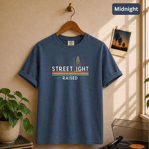 May include: A slate blue short-sleeve t-shirt featuring a graphic of a street light and the text "STREET LIGHT RAISED" in white, positioned above horizontal stripes in orange, yellow, and teal. The shirt is displayed on a wooden hanger.