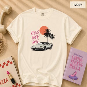 May include: An ivory short-sleeve t-shirt featuring a graphic print of a sports car, two palm trees, and a red sun. The text 'REG BEV WIL' is printed in pink letters to the left of the car. The shirt has a crew neckline and a relaxed fit.