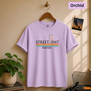 May include: A light purple short-sleeve t-shirt featuring a graphic of a streetlamp and the text "STREETLIGHT RAISED" above horizontal stripes in yellow, orange, teal, and blue. The shirt has a crew neckline and is displayed on a wooden hanger.