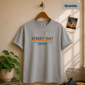 May include: A granite-colored short-sleeve t-shirt featuring a graphic print of a street light and the text STREET LIGHT RAISED. The design includes horizontal stripes in orange, yellow, and blue colors. The shirt has a crew neckline and a standard fit.