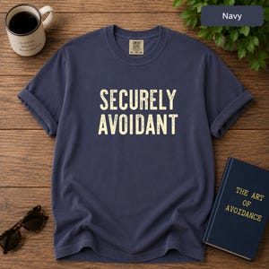 May include: A navy blue short-sleeve t-shirt featuring the text SECURELY AVOIDANT in a distressed cream-colored font on the chest. The shirt has a crew neckline and a Comfort Colors brand label on the inside collar area.