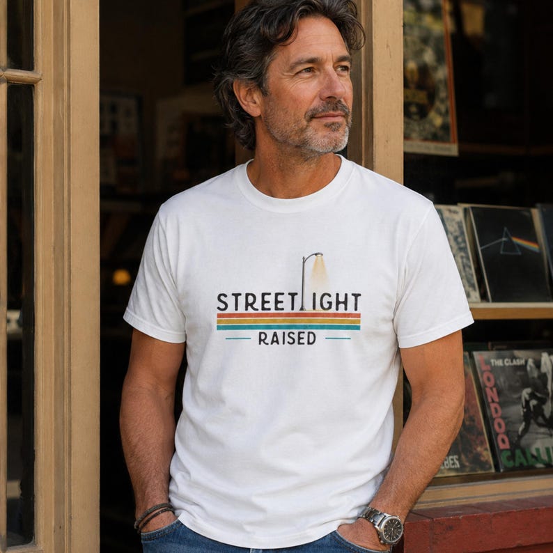 May include: A white short-sleeved t-shirt featuring a graphic print of a street lamp and the text STREET LIGHT RAISED. The design includes horizontal stripes in shades of orange, green, and blue positioned below the word STREET LIGHT.