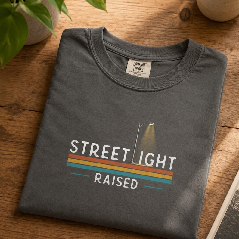 May include: A charcoal gray t-shirt with a graphic print featuring a street lamp and the text STREET LIGHT RAISED. The design includes horizontal stripes in orange, yellow, and teal colors. The shirt has a crew neckline and a Comfort Colors brand label.