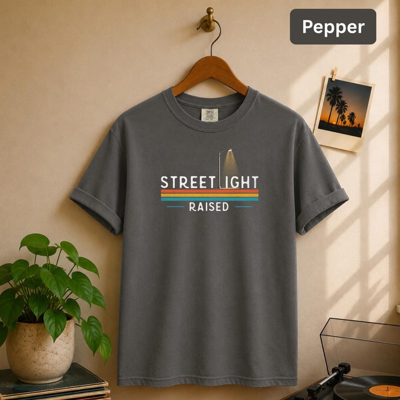 May include: A charcoal gray crew neck t-shirt featuring a graphic print of a street light and the text STREET LIGHT RAISED. The design includes horizontal stripes in orange, yellow, and blue. The shirt is displayed on a wooden hanger.