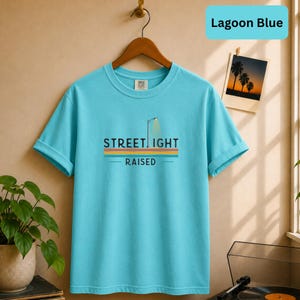 May include: A lagoon blue short-sleeve t-shirt featuring a graphic of a street lamp and the text "STREET LIGHT RAISED" in black, positioned above horizontal stripes in yellow, orange, and teal. The shirt is displayed on a wooden hanger.