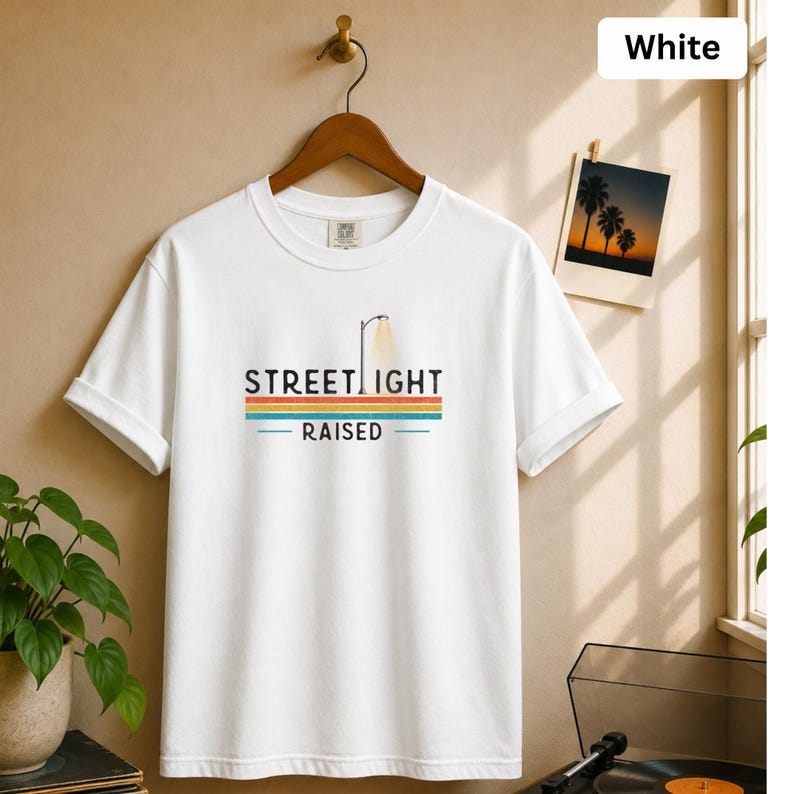 May include: A white short-sleeve t-shirt featuring a graphic print with the text STREET LIGHT RAISED. The design includes a stylized street lamp icon above horizontal stripes in orange, yellow, and blue colors, centered on the chest area.