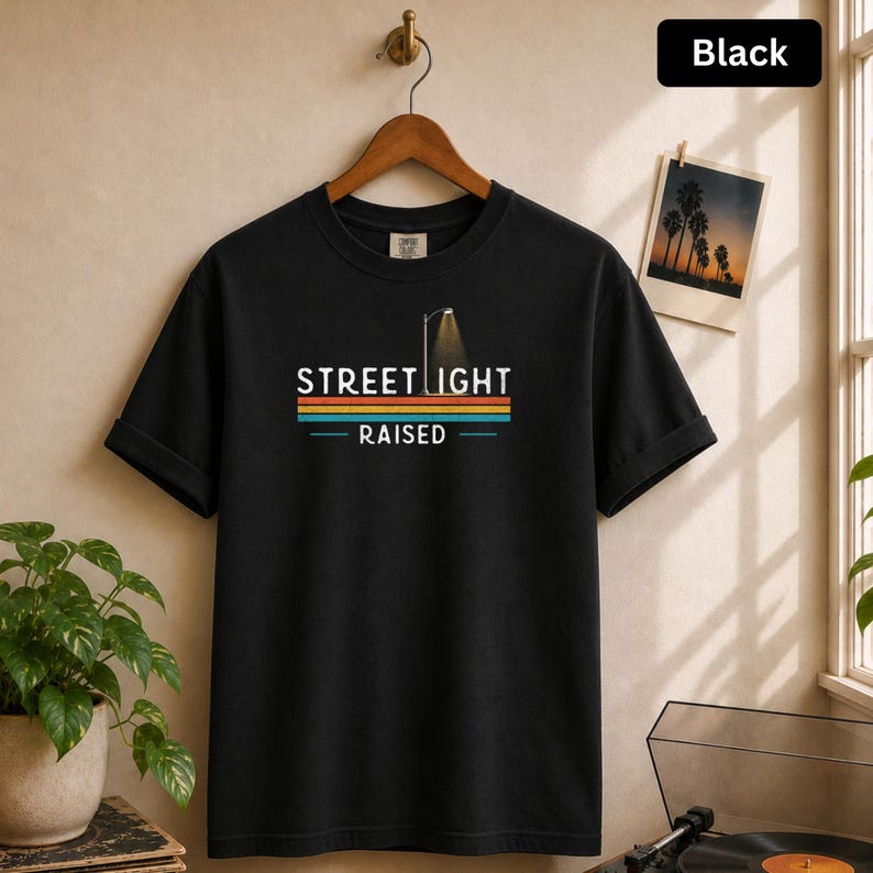 May include: A black short-sleeve t-shirt featuring a graphic of a street light and the text STREET LIGHT RAISED. Below the text are horizontal stripes in orange, teal, and red. The shirt is displayed on a wooden hanger against a neutral wall.