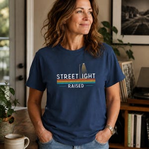 May include: A navy blue short-sleeve t-shirt featuring a graphic of a street light and the text "STREET LIGHT RAISED" in white, positioned above horizontal stripes in shades of orange, yellow, and teal on the center of the chest.