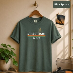 May include: A dark green short-sleeve t-shirt featuring a graphic print with the text STREET LIGHT RAISED. The design includes a street lamp icon and horizontal stripes in orange, yellow, and blue. The shirt is displayed on a wooden hanger.