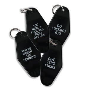 May include: Four black motel-style keychains with silver key rings. Each keychain has white text. The text on the keychains reads: "YOU WERE A FUCK YES FROM DAY ONE", "DO FUCKING YOU", "YOU'RE WORTH THE COMMUTE", and "GIVE ZERO FUCKS".