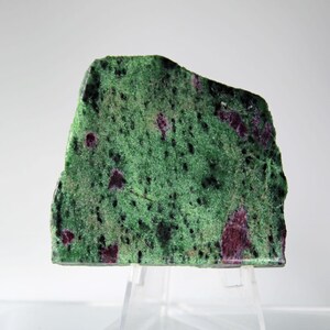 May include: A rectangular slab of green zoisite with deep red ruby inclusions. The stone has a rough, natural edge and is displayed on a clear stand against a white background. The surface is speckled with dark spots.