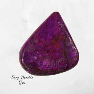 May include: A polished, triangular gemstone with a deep purple hue, speckled with darker shades and hints of red. The stone has a smooth, glossy surface. The text "Stony Mountain Gems" is printed in cursive at the bottom.