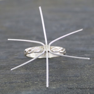May include: Silver ring setting with six prongs, ready to hold a gemstone. The ring band has a braided design.