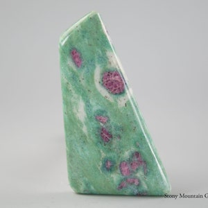 May include: A polished gemstone with a unique shape. The stone features a vibrant green base color with swirling patterns and striking red inclusions. The gemstone is a natural mineral specimen, perfect for collectors or jewelry makers.