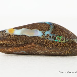 May include: A polished boulder opal gemstone featuring a dark brown, textured ironstone matrix with a horizontal vein of iridescent blue, green, and yellow flashes. The text Stony Mountain Gems is printed in the bottom right corner.