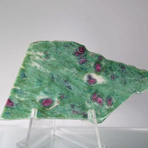 May include: A polished slab of green zoisite with vibrant red ruby inclusions. The stone is irregularly shaped and displayed on a clear acrylic stand, showcasing its natural beauty. The colors are a striking contrast.