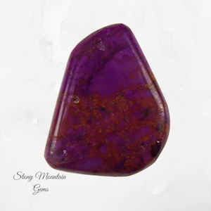 May include: A polished, irregularly shaped gemstone with a deep purple hue, accented by reddish-brown veining. The stone has a glossy finish and the text "Stony Mountain Gems" is visible.