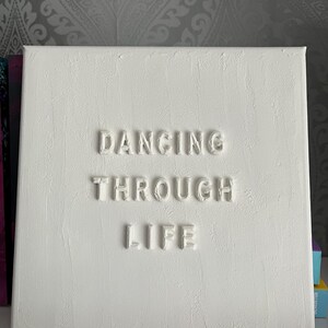 Op de afbeelding: Wit canvas met de zin "Dancing Through Life" in reli&euml;f in witte letters.