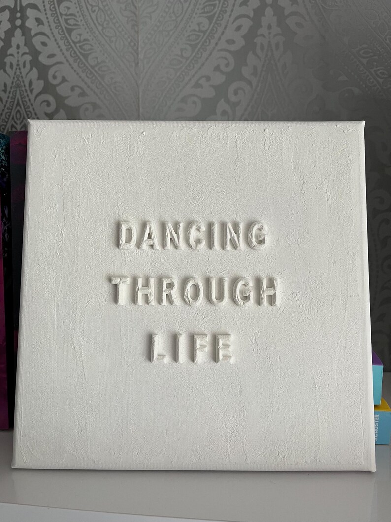 Op de afbeelding: Wit canvas met de zin "Dancing Through Life" in reli&euml;f in witte letters.