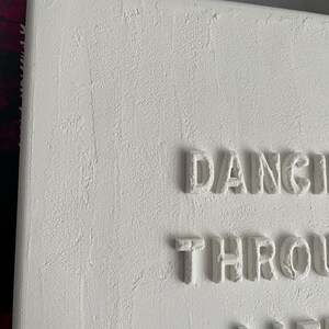 Op de afbeelding: Witte canvas wandkunst met de woorden "Dancing Through Life" in reli&euml;fletters.