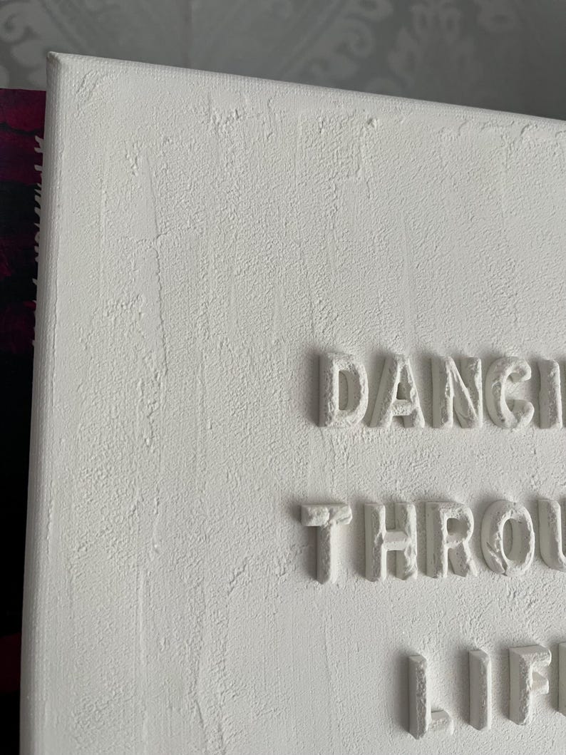 Op de afbeelding: Witte canvas wandkunst met de woorden "Dancing Through Life" in reli&euml;fletters.