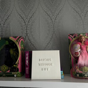 Op de afbeelding: Drie dozen op een wit rek met een grijze, gepatroneerde achtergrond. De doos links heeft een zwarte en groene Wicked Witch of the West pop. De middelste doos is wit met de tekst "Dancing Through Life" in reli&euml;f op de voorkant. De doos rechts heeft een roze en witte Wicked Witch of the West pop.