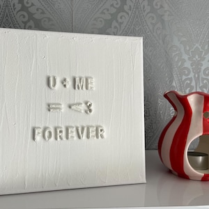 May include: A square white canvas featuring raised white lettering that reads U + ME = <3 FOREVER. The surface of the canvas has a textured, vertical brushstroke finish. Beside the canvas sits a ceramic wax warmer with red and white vertical stripes.