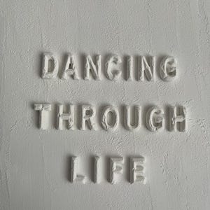 Op de afbeelding: Witte muur met de woorden "Dancing Through Life" in reli&euml;f.