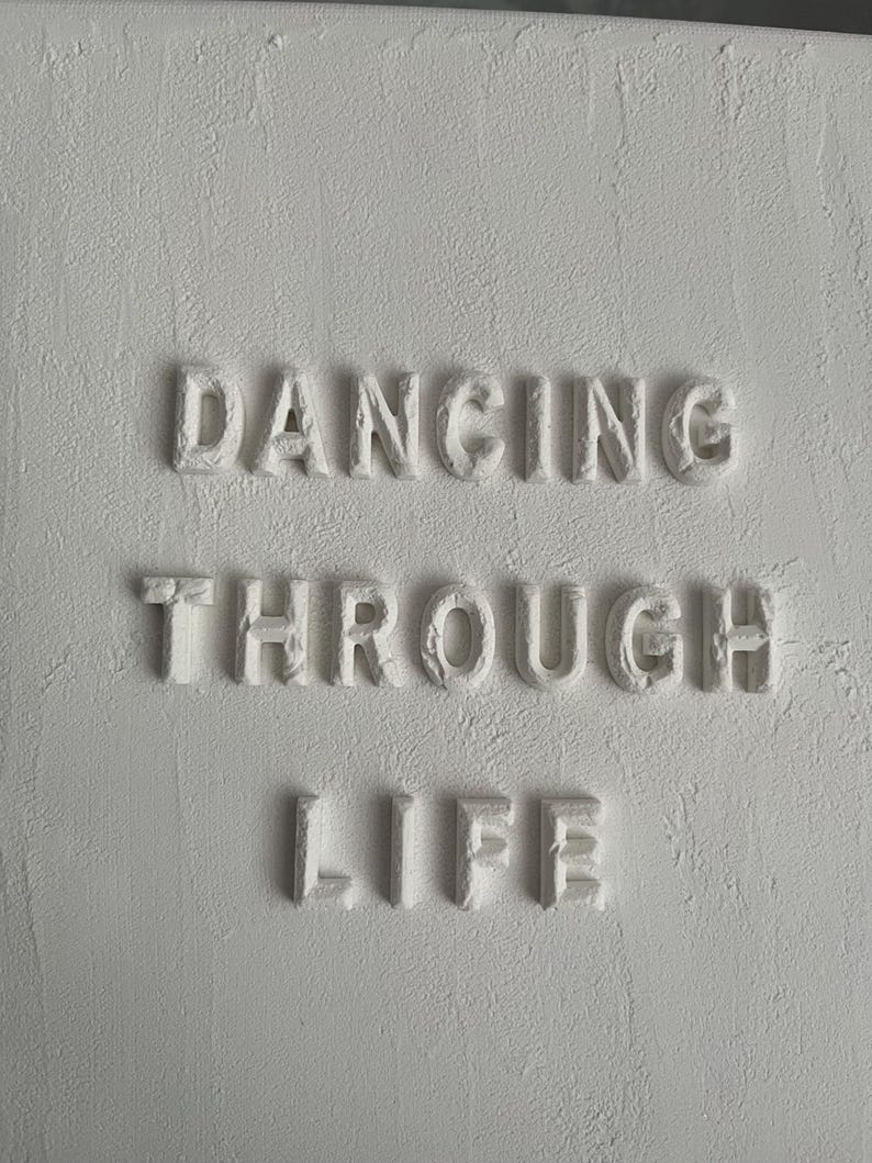 Op de afbeelding: Witte muur met de woorden "Dancing Through Life" in reli&euml;f.