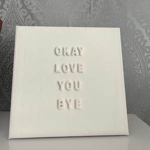 May include: A square, white canvas with the words "OKAY LOVE YOU BYE" in raised, off-white letters. The canvas is on a white surface, with a wooden picture frame and sunglasses visible in the background.