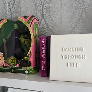 Op de afbeelding: Een roze en groene doos met een pop van de Wicked Witch of the West uit The Wizard of Oz. De doos is gelabeld met "Wicked" en "Deluxe Edition". Twee boeken staan op de plank achter de doos, een is roze en heet "Say You Swear" van Meagan Brandy, de andere is wit en heet "Dancing Through Life".