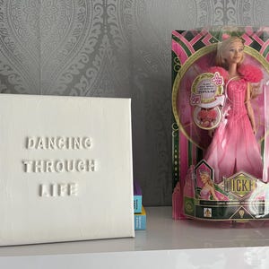Op de afbeelding: Wit canvas met de woorden "Dancing Through Life" in reli&euml;f. Een pop in een roze jurk en een kroon zit in een doos achter het canvas.