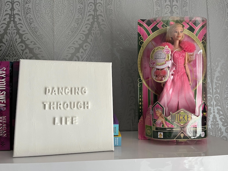 Op de afbeelding: Wit canvas met de woorden "Dancing Through Life" in reli&euml;f. Een pop in een roze jurk en een kroon zit in een doos achter het canvas.