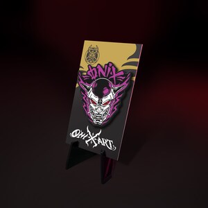 May include: A black and white Oni mask enamel pin with purple accents and red eyes, displayed on a black card with the text "ONIX ART" and a logo. The pin is set against a gradient background.
