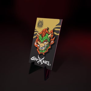 May include: A colorful enamel pin featuring a green and orange Bowser design. The pin is mounted on a black and gold backing card with the text "ONYX ART" in white. The pin is displayed on a black stand.