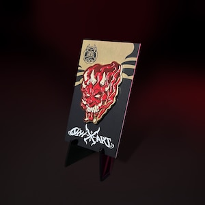 May include: A red and gold enamel pin featuring a demonic oni mask design. The pin is displayed on a black card with the text "ONIX ART" and a small logo. The pin has a gold outline and is set against a dark background.