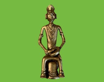 May include: A metallic bronze-toned figurine of a seated person wearing a headpiece and layered necklaces. The figure is depicted in a seated position, holding a circular object in their hands, with a textured surface and a dark, weathered finish.