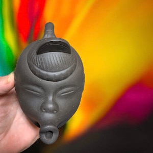 May include: A black ceramic teapot shaped like a human face with a spout and a lid. The teapot has a simple design with closed eyes and a small nose.