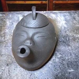 May include: A black ceramic teapot shaped like a face with a spout that looks like a mouth. The teapot has a lid with a textured design.