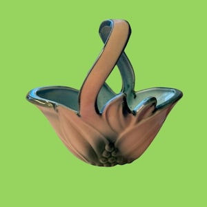 May include: A ceramic basket with a sculptural, flower-like design featuring a high, arched handle. The exterior displays a gradient of soft pink and muted green tones, while the interior is finished in a smooth, light blue glaze.